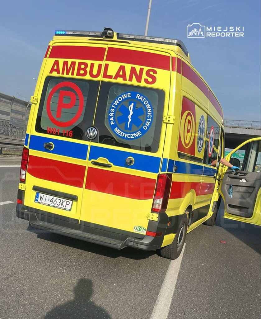 Ambulans z widocznymi oznaczeniami, zaparkowany na pasie drogowym, z otwartymi drzwiami po stronie kierowcy.