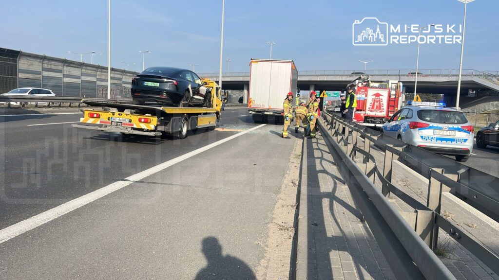 Strażacy i policjanci zabezpieczają miejsce wypadku na drodze, obok lawety z uszkodzonym samochodem.