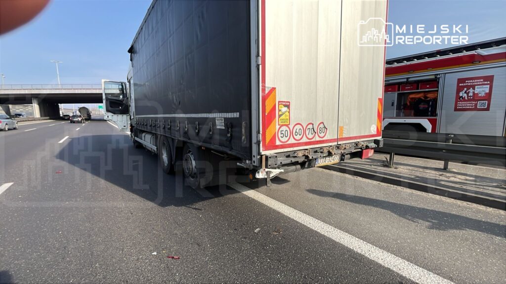 Czarny ciężarówka z otwartymi drzwiami stoi na pasie ruchu obok autostrady, w tle widoczny wóz strażacki.