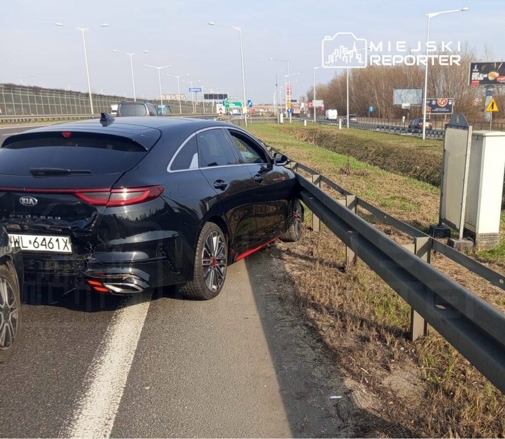 Czarny samochód marki Kia zaparkowany wzdłuż drogi, przy barierce ochronnej, z widocznymi uszkodzeniami tylnej części.