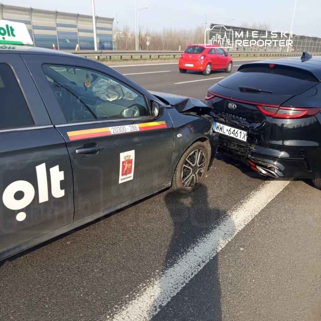 Dwa uszkodzone samochody, taksówka z logo "olt" i czarny pojazd, stoją na pasie drogowym po kolizji.