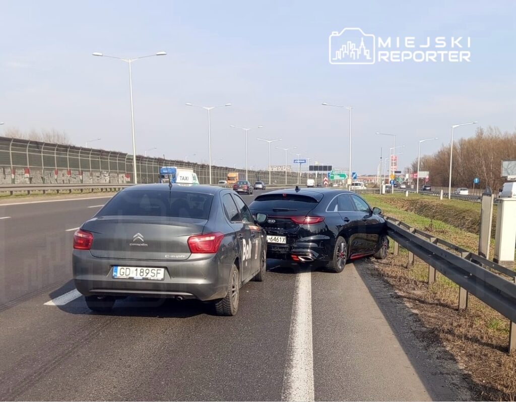 Dwa samochody osobowe, w tym taksówka, stoją na poboczu ruchliwej drogi, obok barierek ochronnych.