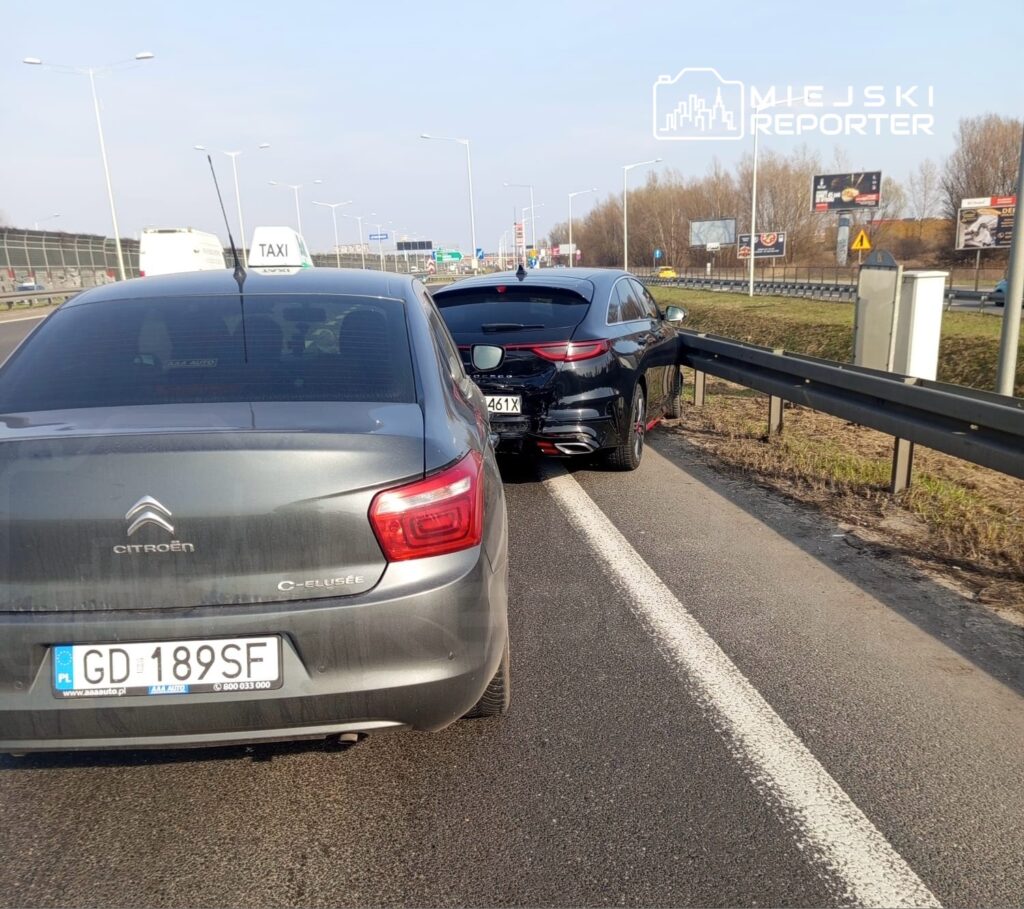 Dwa samochody stoją na pasie awaryjnym drogi, jeden to taksówka, a drugi to czarny pojazd z uszkodzeniami.