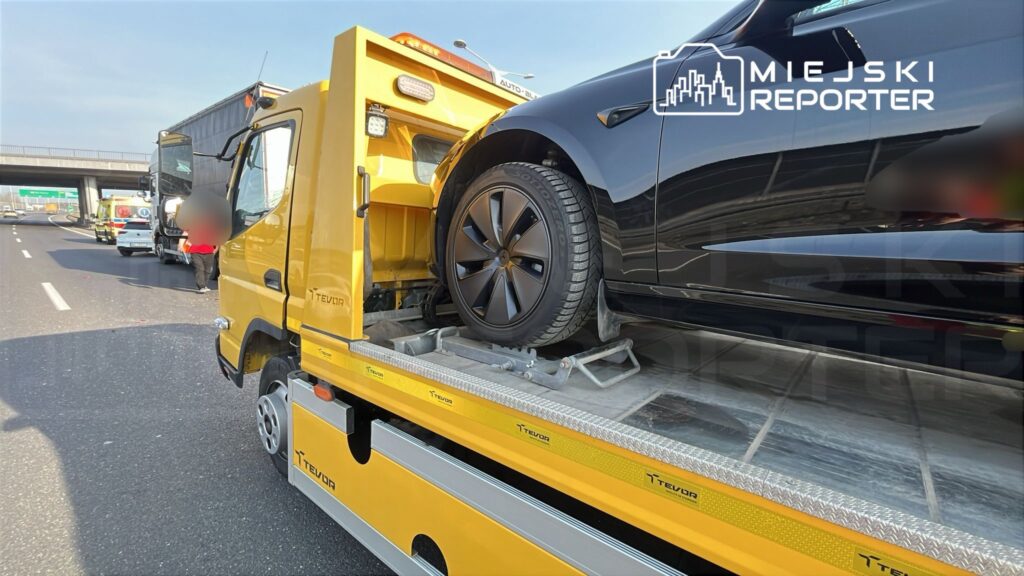 Żółty samochód holowniczy transportuje czarny pojazd osobowy na autostradzie, w tle widoczne inne pojazdy.