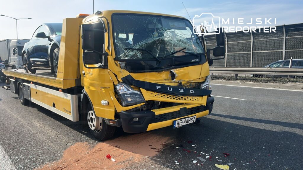 Żółty samochód lawetowy z uszkodzoną przednią częścią transportuje czarny pojazd na autostradzie.