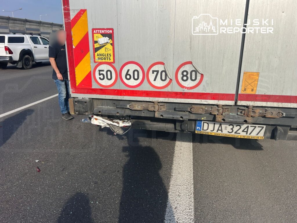 Mężczyzna w niebieskich dżinsach stoi obok tyłu ciężarówki na ruchliwej drodze, z widocznymi znakami prędkości.