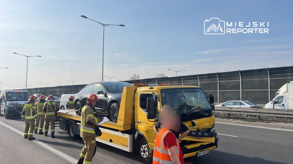 Grupa strażaków w odzieży ochronnej pracuje przy wypadku drogowym na autostradzie, obok holownika z uszkodzonym samochodem.
