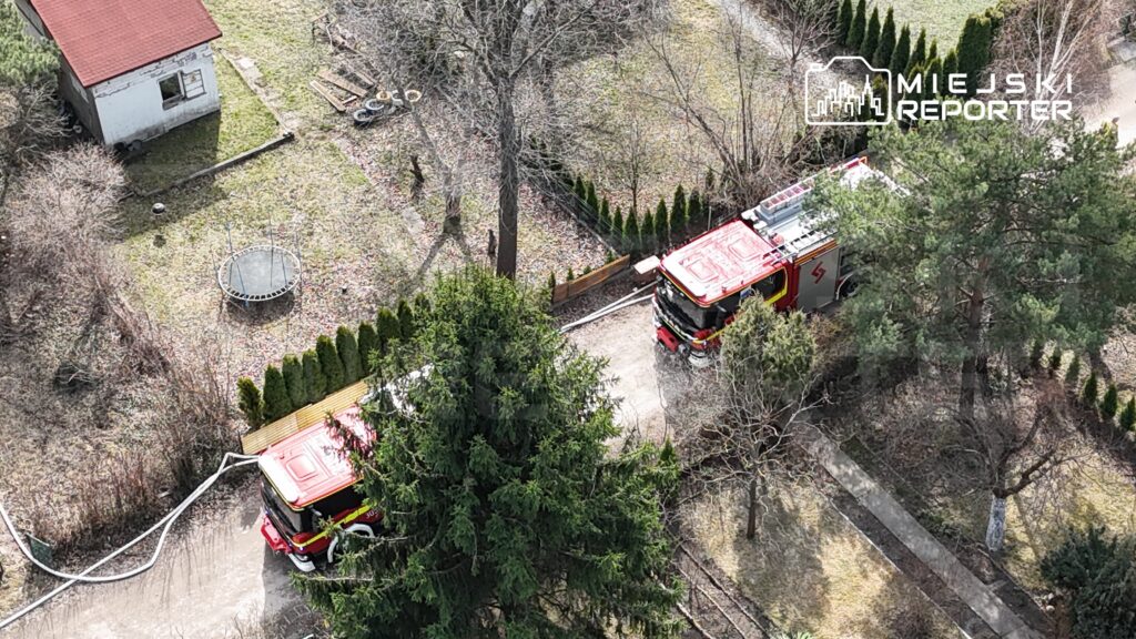 Dwa wozy strażackie stoją na podjeździe, w tle widoczny skokowy trampolina i zarośla.