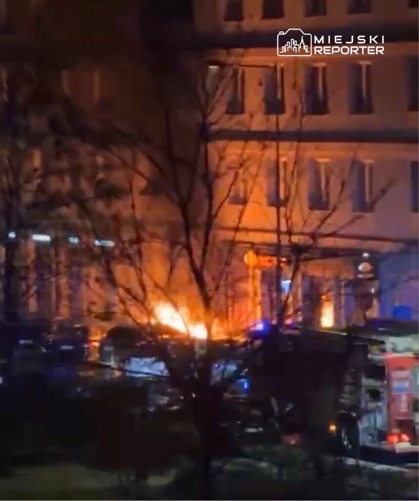 W tle widoczne płomienie wydobywające się z budynku, obok straż pożarna z włączonymi sygnalizatorami świetlnymi.