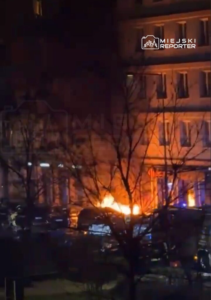 Pojazd płonący na parkingu w nocy, otoczony przez gałęzie drzew i budynki w tle.