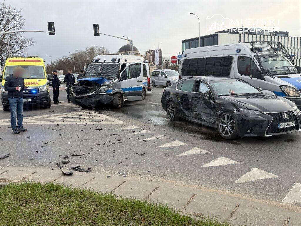 Na skrzyżowaniu leży rozbite auto osobowe i policyjny van, wokół fragmenty pojazdów. Obok stoi mężczyzna z telefonem.