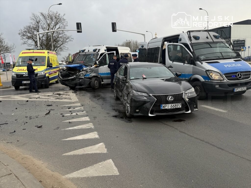 Na skrzyżowaniu stoi uszkodzony samochód osobowy i policyjny furgon, wokół nich pracują funkcjonariusze oraz straż pożarna.