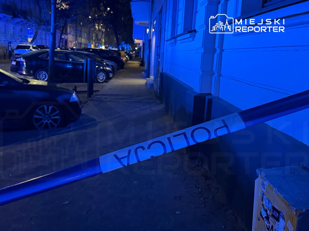 Niebieska taśma policyjna zabezpiecza chodnik w miejskiej okolicy, w tle zaparkowane samochody i oświetlone budynki.