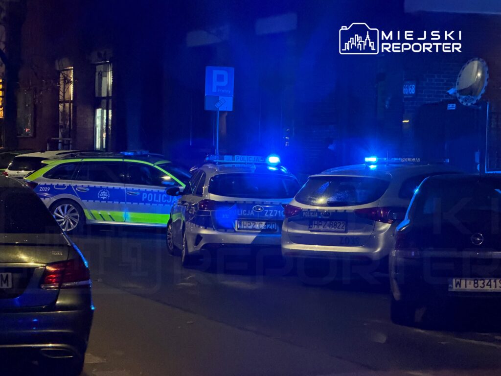 Policyjne samochody z włączonymi sygnałami świetlnymi zaparkowane na ulicy w nocy, w tle widoczne budynki.