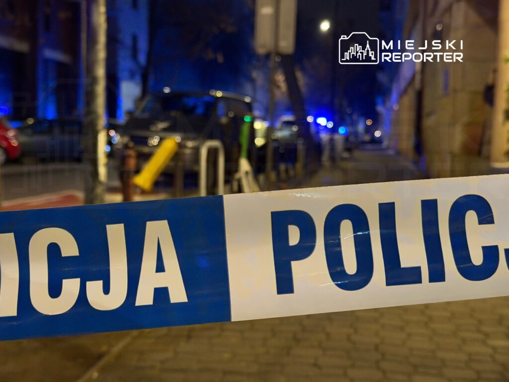 Niebieska taśma policyjna zabezpiecza teren, na którym widać zaparkowane pojazdy oraz oświetlenie w nocy.