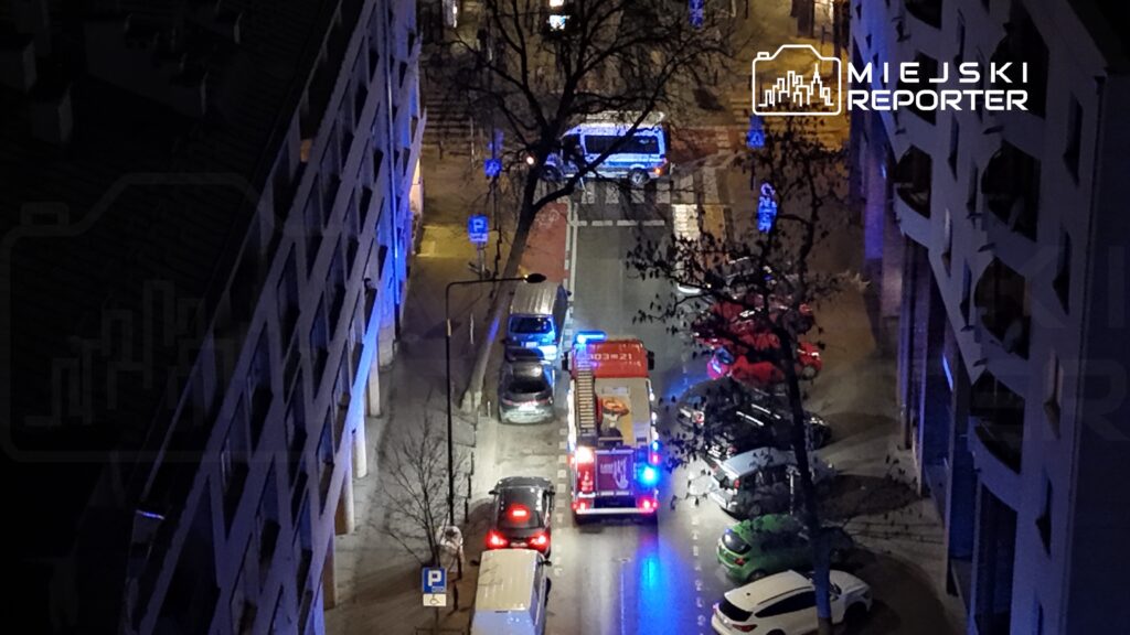 W nocy na ulicy widać straż pożarną z włączonymi sygnałami świetlnymi, otoczoną zaparkowanymi samochodami.