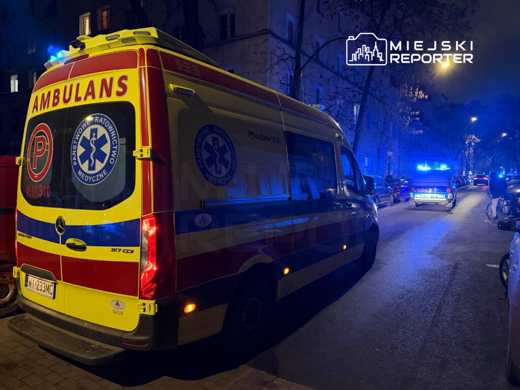 Ambulans z włączonymi sygnałami świetlnymi parkuje na ulicy, obok policyjnego radiowozu z niebieskimi światłami.