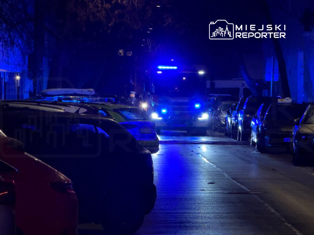 Na ulicy z zaparkowanymi samochodami widoczny jest radiowóz oraz woz strażacki z włączonymi niebieskimi światłami.