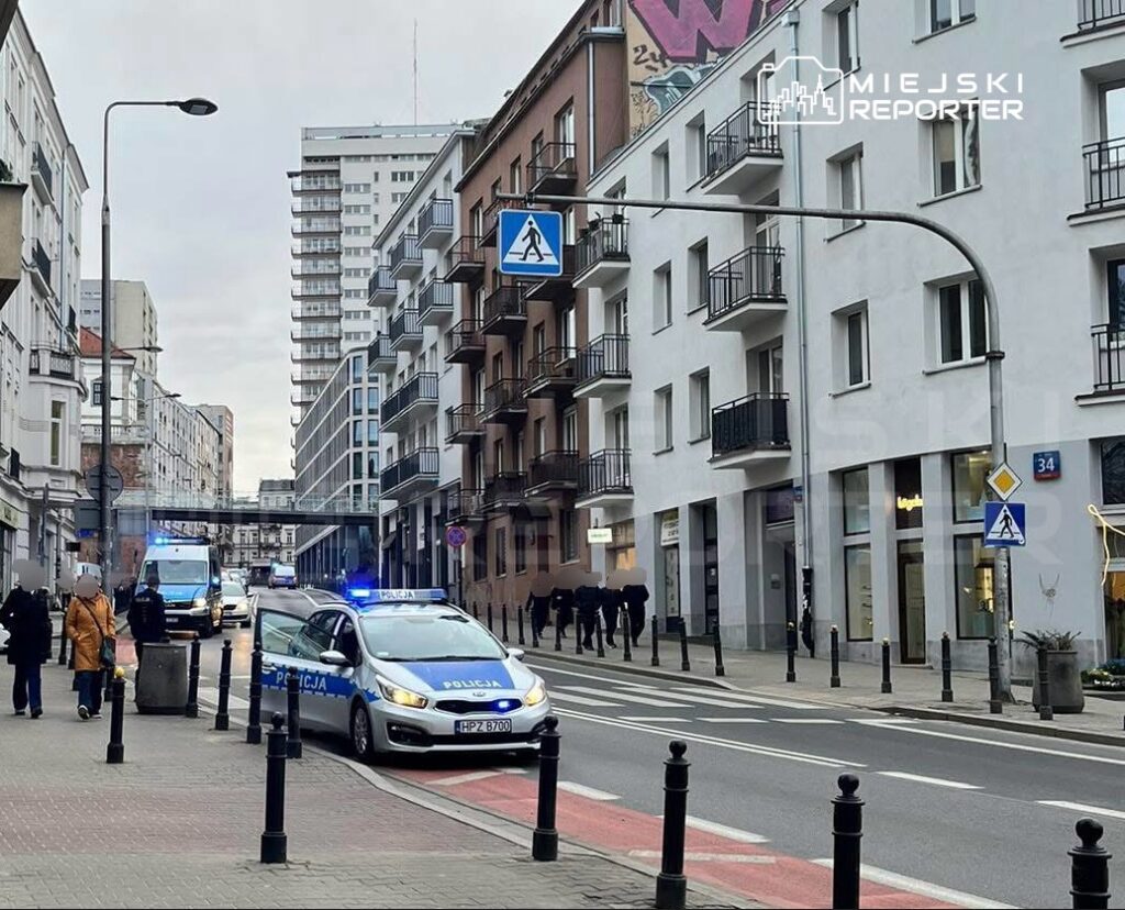 Policyjny radiowóz z włączonymi sygnalizatorami na ulicy, obok przechodniów i nowoczesnych budynków.