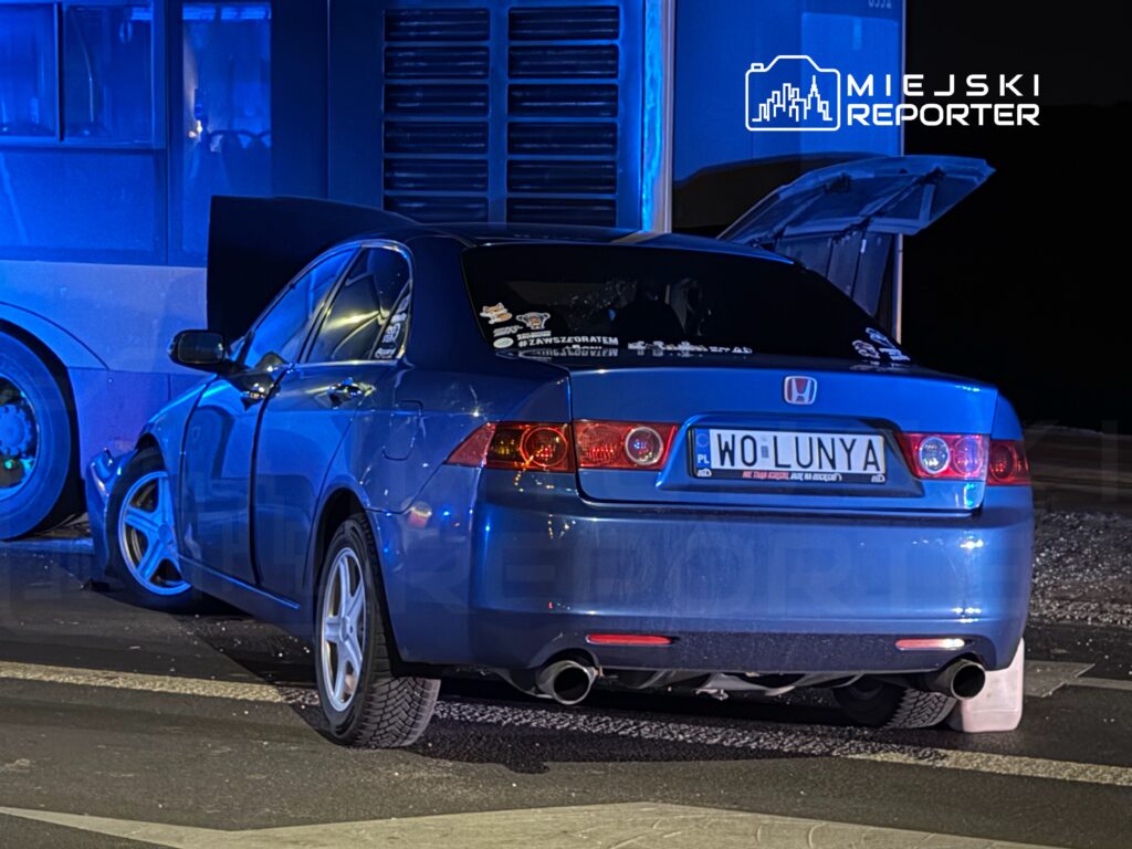 Niebieski samochód osobowy z otwartą klapą bagażnika stoi na drodze w pobliżu przystanku autobusowego, oświetlony niebieskim światłem.