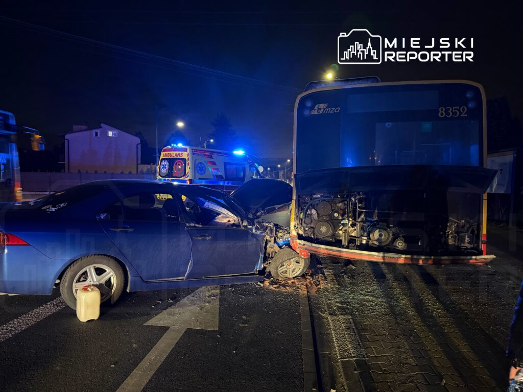 Niebieski samochód osobowy z uszkodzoną boczną częścią stoi obok przystanku, a w tle widoczny jest autobus oraz karetka pogotowia.