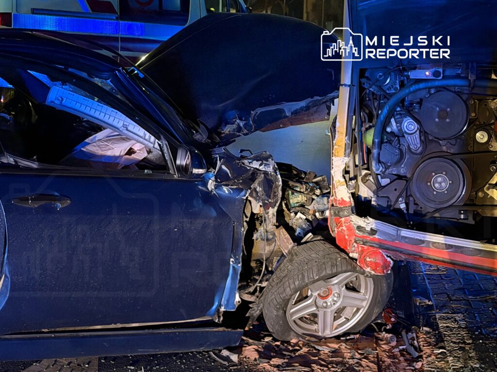 Zniszczony niebieski samochód osobowy z wgniecioną stroną, stojący obok uszkodzonego pojazdu, w tle świecące światła służb ratunkowych.