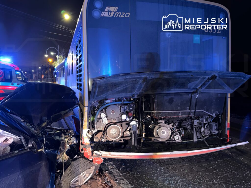 Czarny samochód osobowy z uszkodzoną przednią częścią zderza się z autobusem, w tle widać pojazdy ratunkowe.