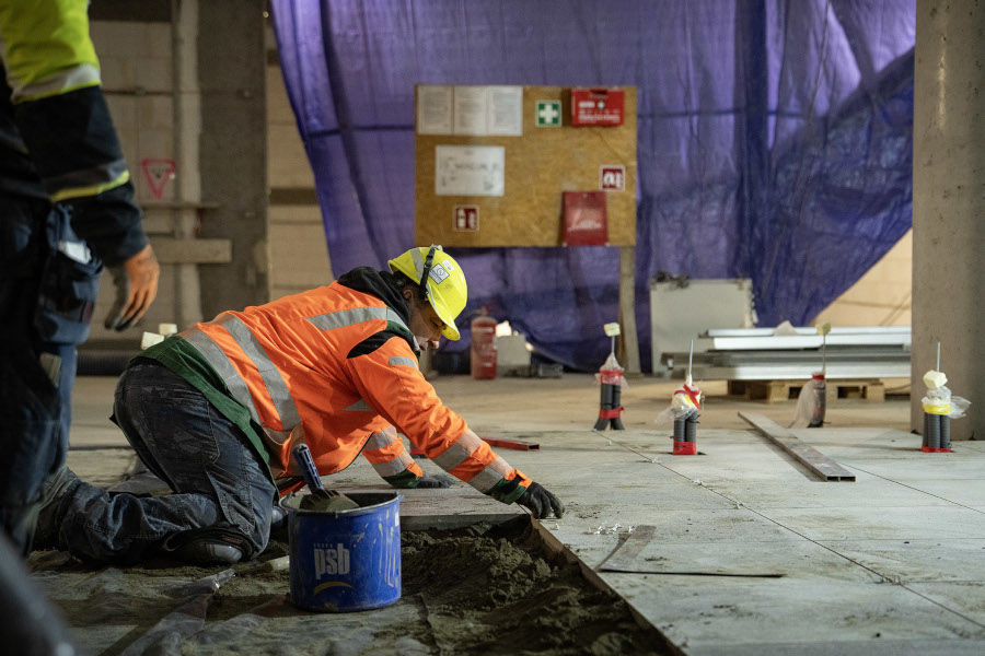 Robotnik w pomarańczowej kamizelce układa podłogę w budynku w trakcie budowy, otoczony sprzętem i materiałami budowlanymi.