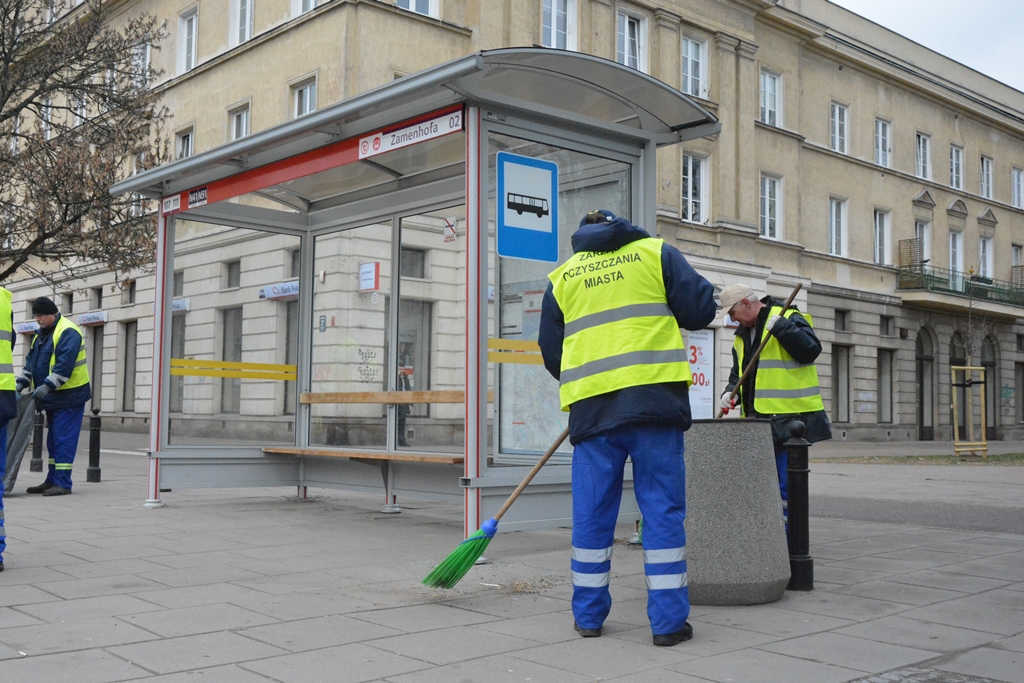 Pracownicy służb porządkowych sprzątają przystanek autobusowy, używając miotły i narzędzi, w miejskim otoczeniu.