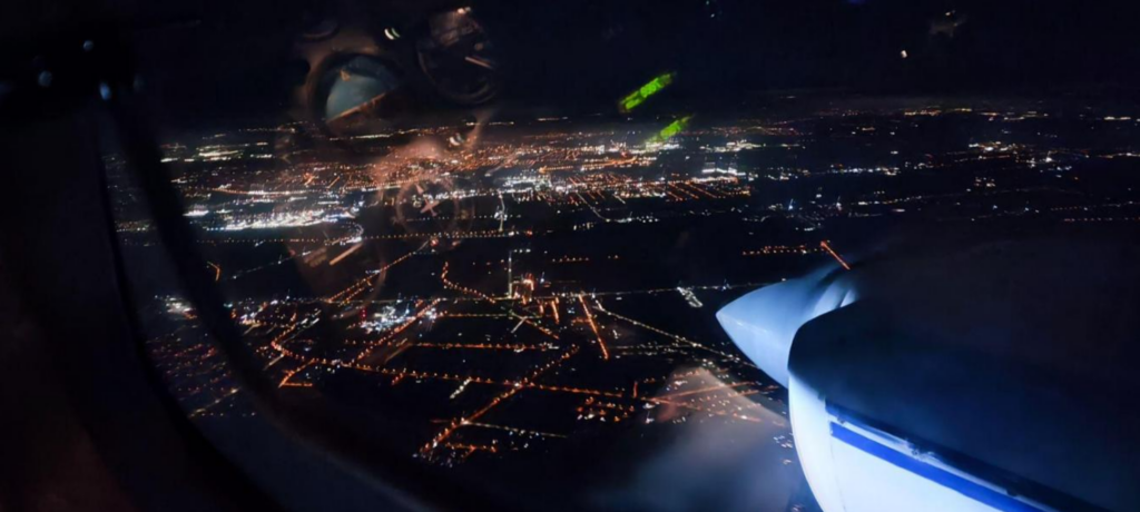 Widok z okna samolotu na nocne oświetlenie miasta, z widoczną śmigłową częścią silnika.