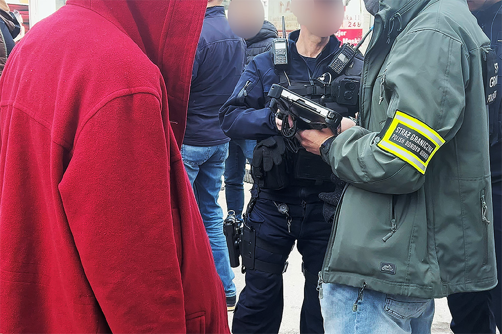 Mężczyzna w czerwonym kapturze stoi obok funkcjonariusza Straży Granicznej z bronią w ręku, na tle grupy ludzi.