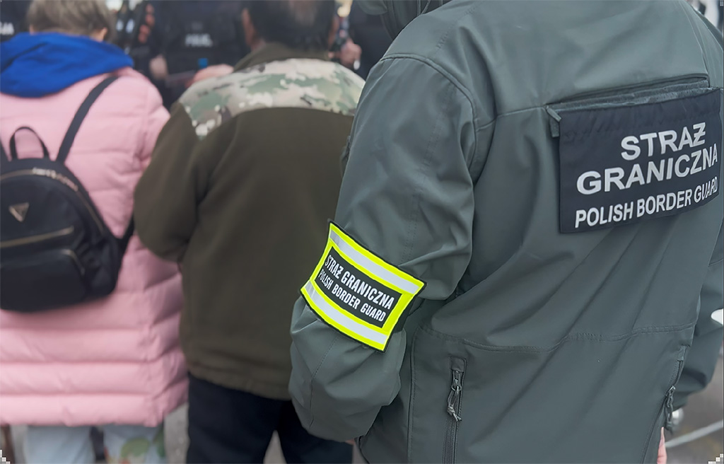 Funkcjonariusz Straży Granicznej z oznaczeniem "POLISH BORDER GUARD" stojący w tłumie ludzi.