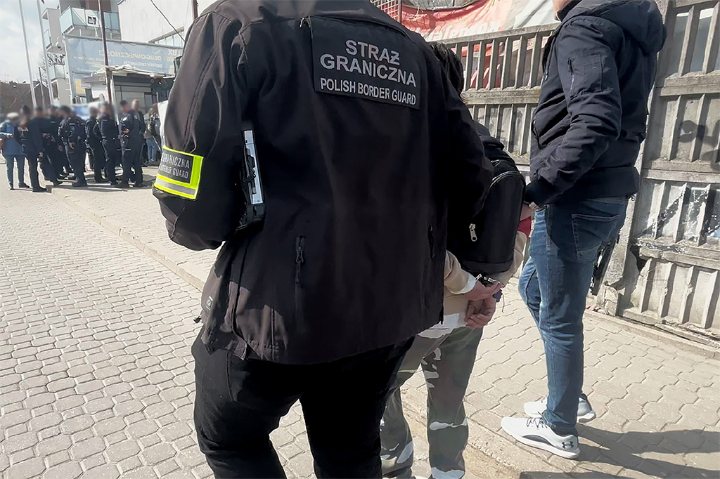 Funkcjonariusz Straży Granicznej zatrzymuje osobę w okolicy przejścia granicznego, w tle grupa policjantów.