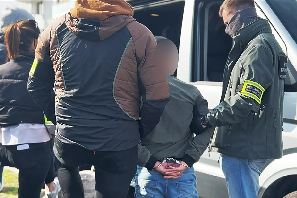 Grupa funkcjonariuszy zatrzymuje osobę z rękami za plecami w pobliżu białego pojazdu.