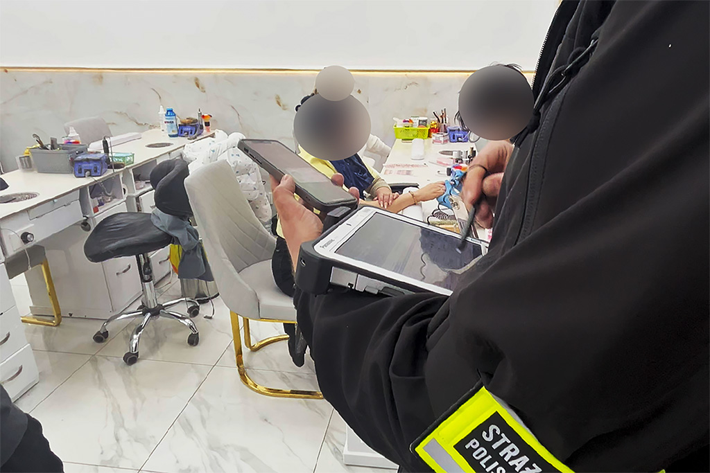 Funkcjonariusz straży miejskiej korzysta z urządzenia mobilnego w salonie kosmetycznym, w tle klienci przy stolikach.