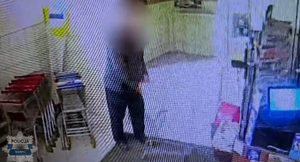 Mężczyzna w szarej bluzie stoi w pomieszczeniu z wózkami na zakupy w tle.