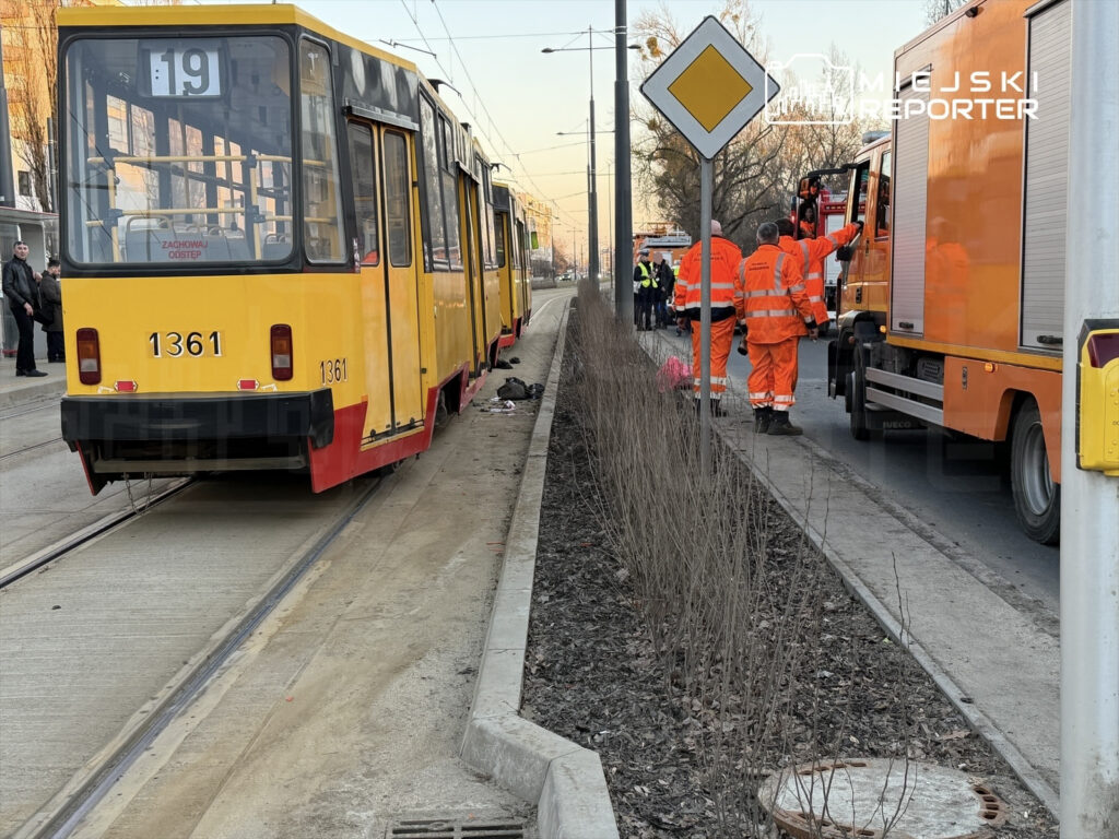 Tramwaj linii 19 zatrzymany na przystanku, obok pracownicy w pomarańczowych uniformach prowadzący prace na torach.