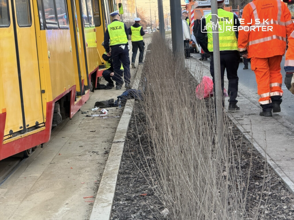 Policjanci oraz ratownicy medyczni udzielają pomocy osobie leżącej obok tramwaju na przystanku.