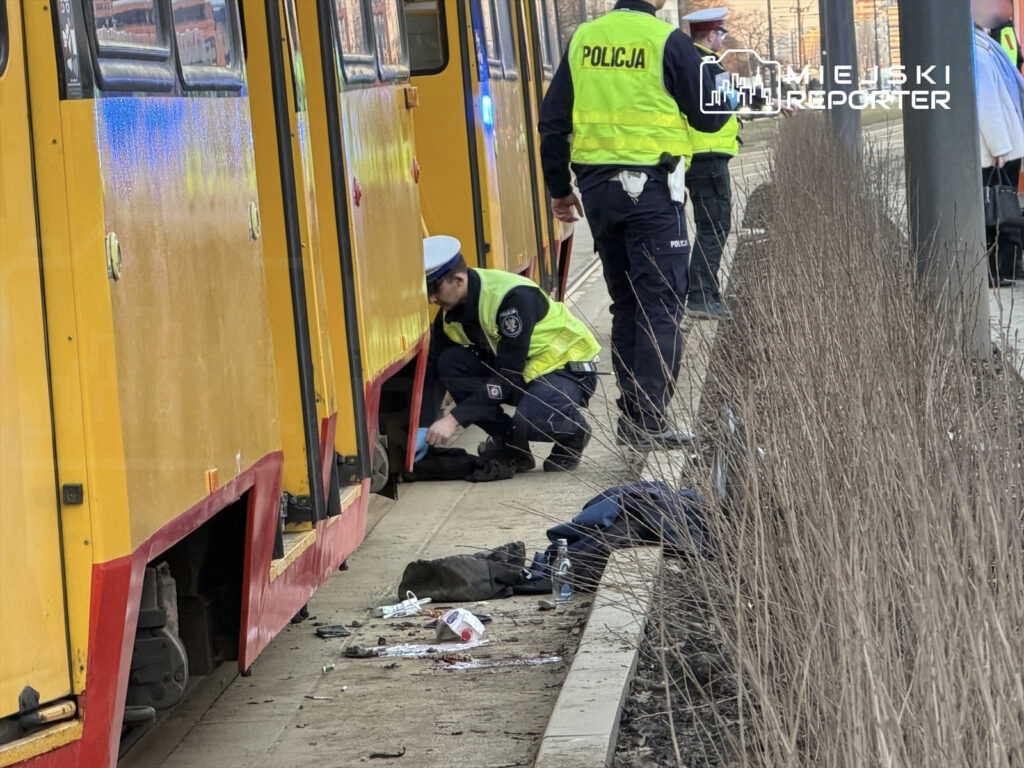 Policjanci w kamizelkach odblaskowych sprawdzają osobę leżącą obok żółtego tramwaju na przystanku.