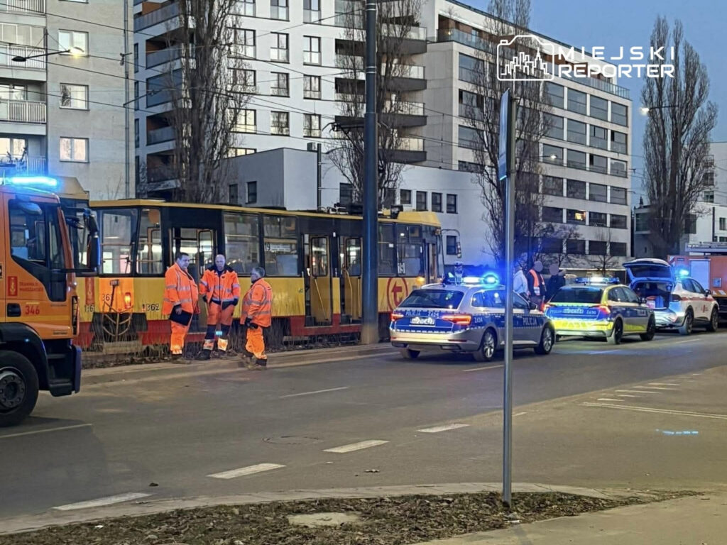 Grupa pracowników w pomarańczowych kombinezonach naprawia tramwaj obok zaparkowanych radiowozów policyjnych.
