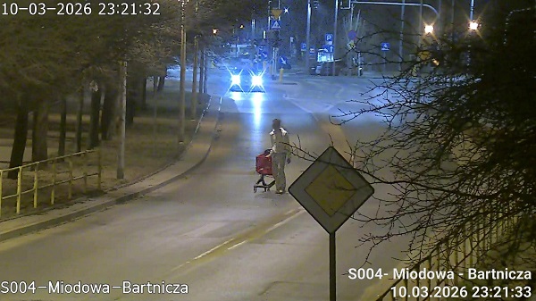 Mężczyzna z wózkiem na zakupy przechodzi przez pustą ulicę w nocy, w tle widać samochody i latarnie.