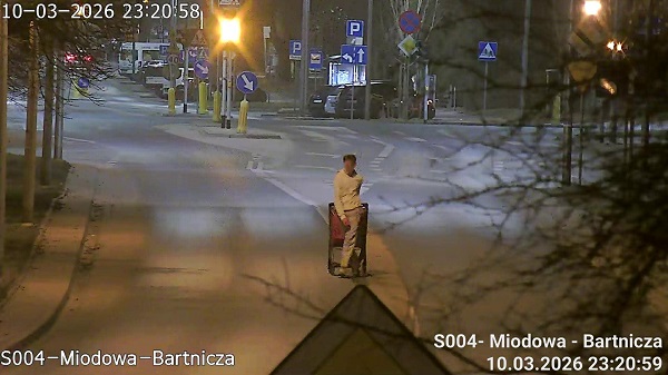 Mężczyzna w białej odzieży stoi z wózkiem na oświetlonej ulicy, w tle widoczne znaki drogowe i zaparkowane samochody.