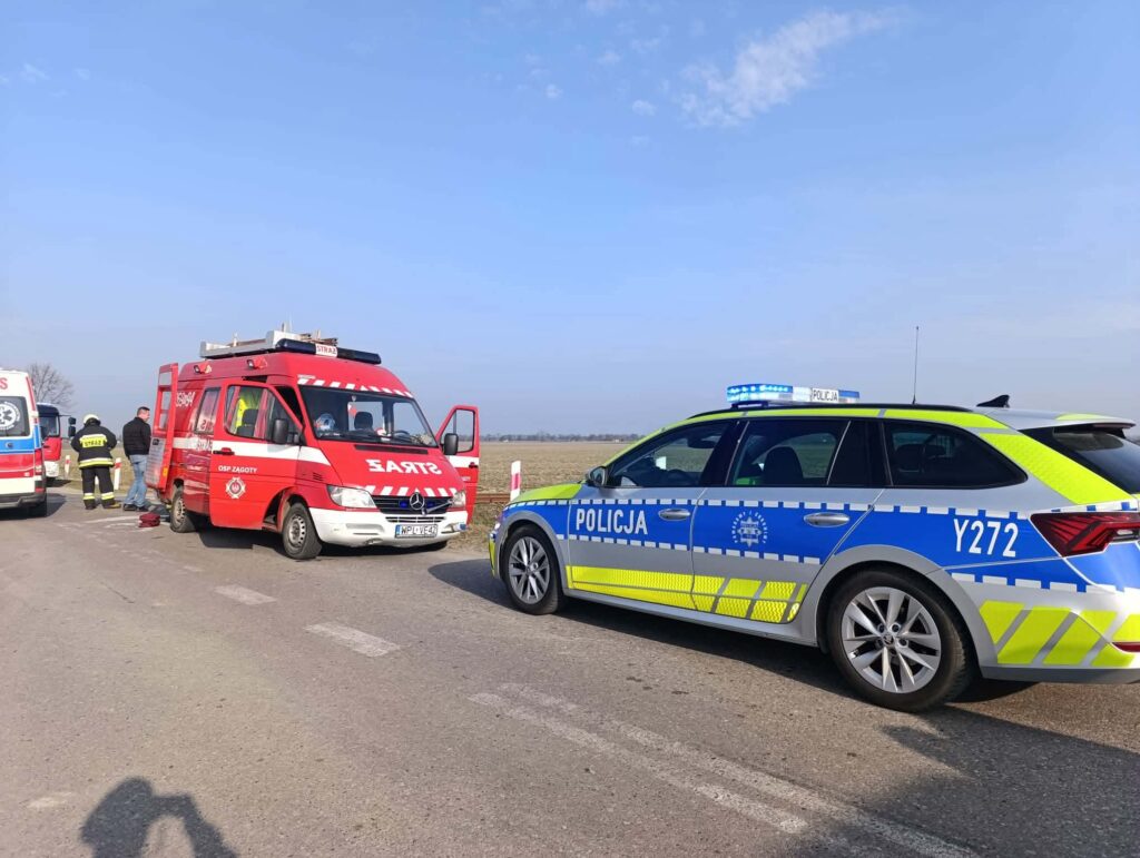 Na drodze zatrzymane są pojazdy służb ratunkowych, w tym karetka i radiowóz policji, obok stoją strażacy.
