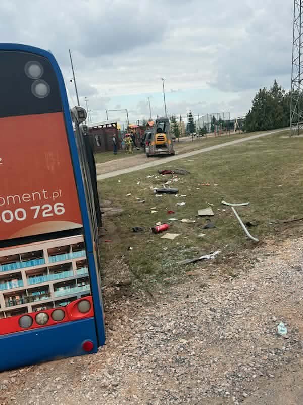 Przewrócony autobus leży na boku na trawiastej przestrzeni, wokół porozrzucane śmieci i sprzęt budowlany.