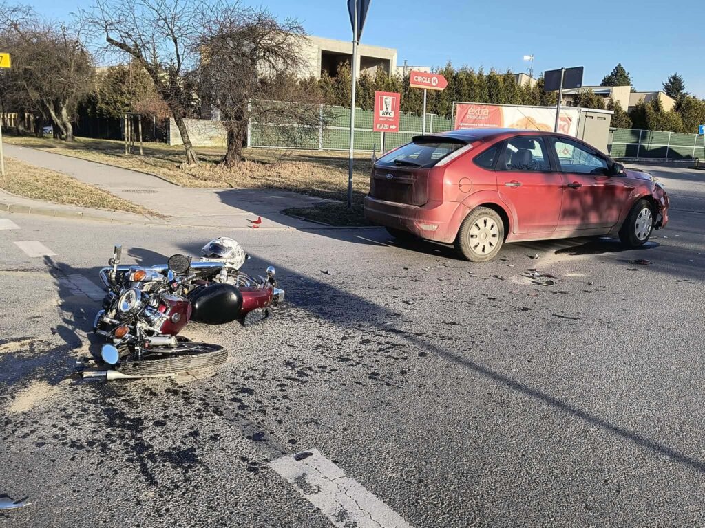 Motocykl leży na jezdni, obok uszkodzony czerwony samochód osobowy, w tle widoczna zieleń i reklamy.