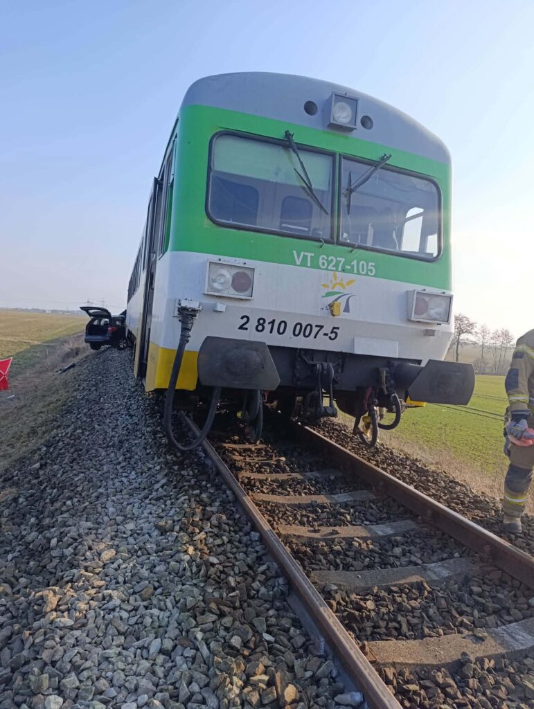 Pociąg z zielonymi detalami stoi na torach, obok pracuje strażak z narzędziami. W tle rozciągają się pola.