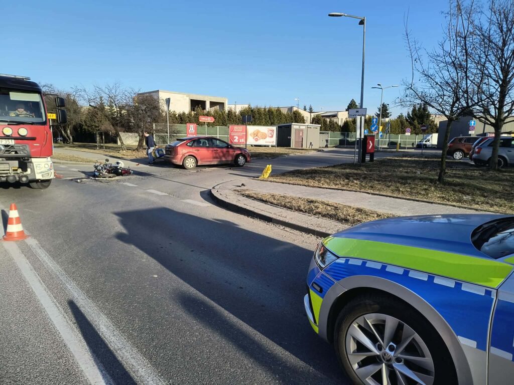 Wypadek drogowy z udziałem samochodu osobowego i motocykla na skrzyżowaniu, obok stojącego wozu strażackiego.