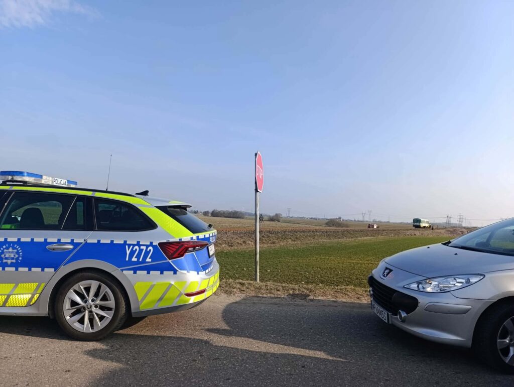 Policyjny radiowóz z niebieskimi i żółtymi oznaczeniami stoi przy znaku stop na wiejskiej drodze. Obok zaparkowany srebrny samochód osobowy.