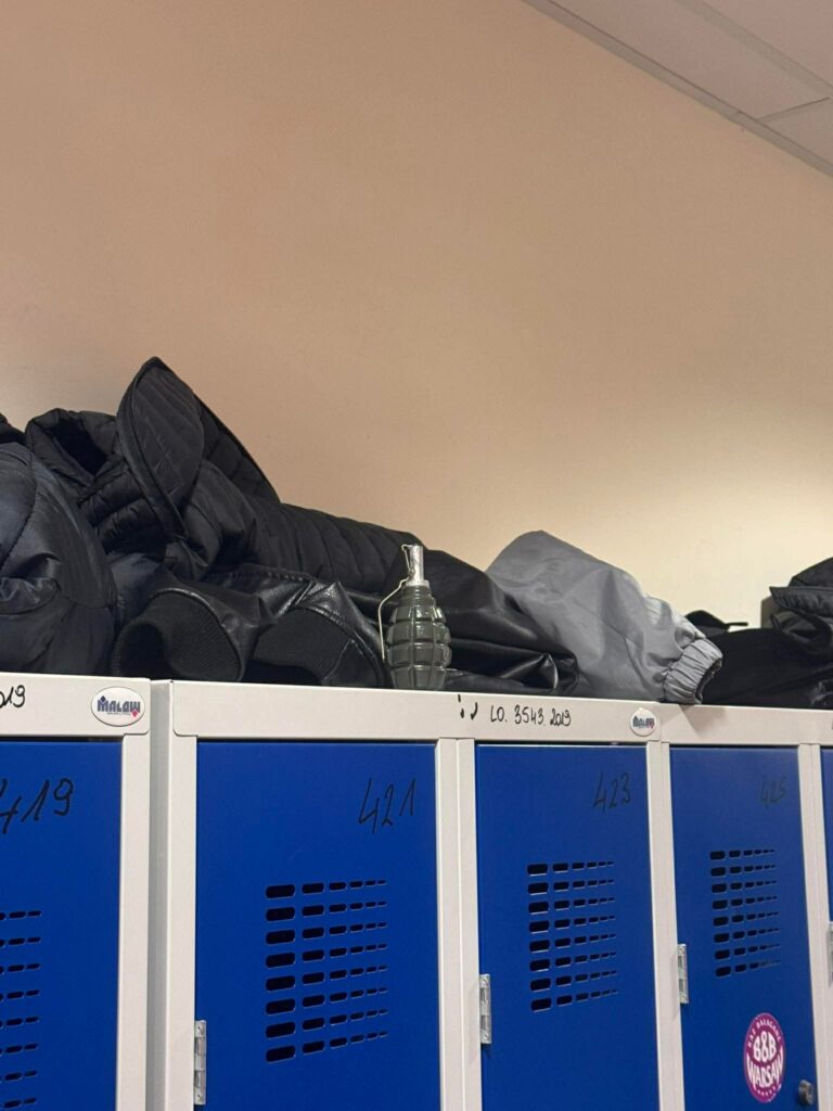 Na górze niebieskich szafek leżą czarne kurtki, a obok nich znajduje się plastikowa butelka w kształcie granatu.