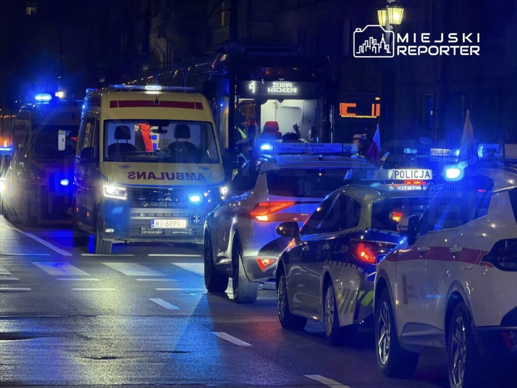 Karetka pogotowia oraz radiowozy policyjne zatrzymane na oświetlonej ulicy nocą, otoczone przez niebieskie światła.